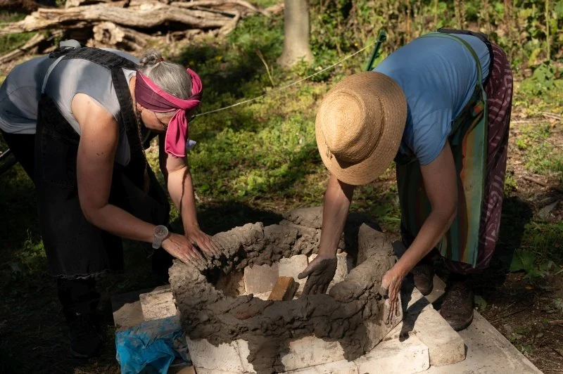 Two women are building an outdoor kiln out of clay in the sunshine