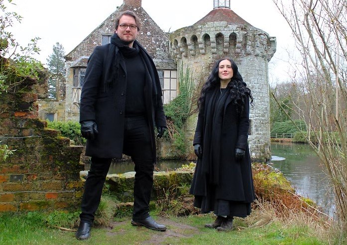 A man and a woman stand in front of a small gothic style building by a river