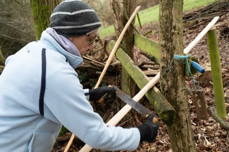 A woman carving a tree branch into a smooth post using a hand saw