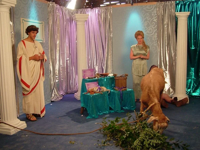 Two people dressed as ancient greeks with goat in foreground
