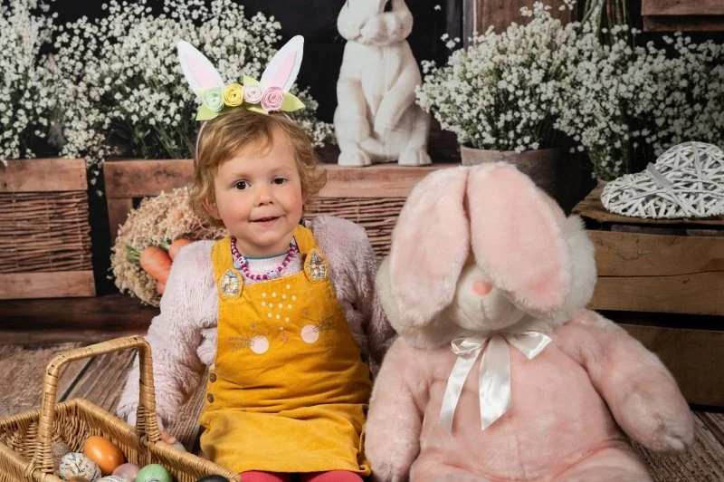 A smiling girl sitting next to a large pink bunny soft toy in an Easter themed photo studio