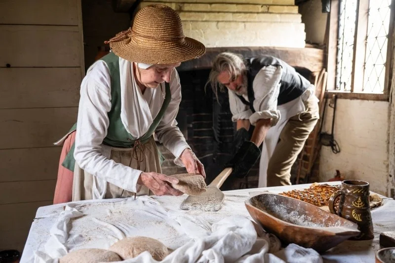 A woman dressed in 18th century clothing with a straw hat places a shaped piece of dough onto a wooden padle so that the man standing next to her can place it into a traditional oven in the 18th century cottage they are both standing in
