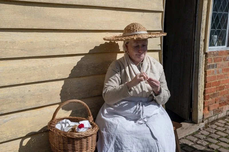 A lady dressed in 18th century style clothing and a straw hat makes buttons using scraps of material from her wicker basket.