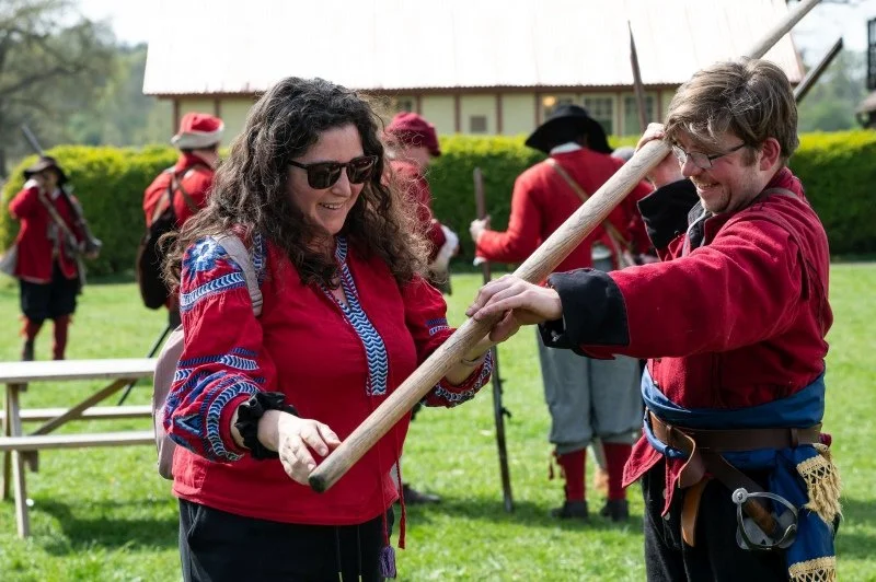 A lady with long brown hair is being handed a long wooden pike by an English Civil War reenactor in costume