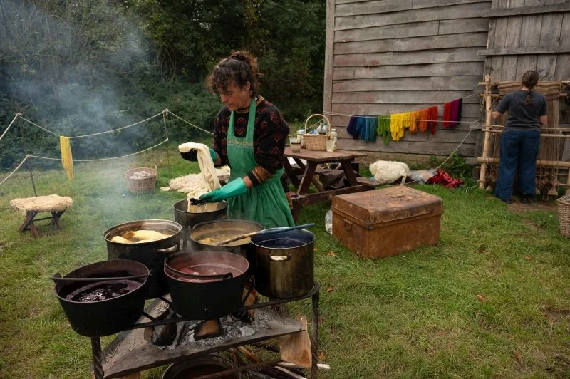 A woman stands behind a group of pots holding different coloured dyes while another woman weaves wool on a traditional loom in the background