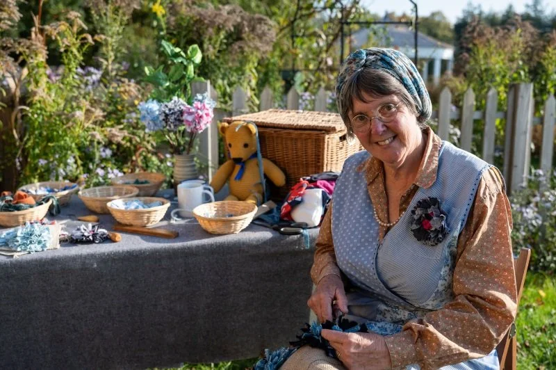 A woman dressed in 1940s style cloths sits by a table filled with rag rug scraps and tools