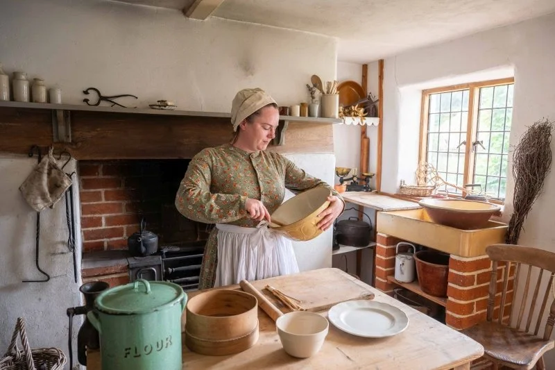 A woman dressed in victorian clothing is cooking in the kitchen of a cottage