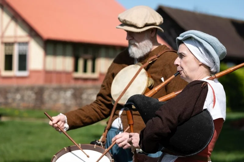 A lady dressed in Tudor period dress plays the bagpipes while a man in a similar themed clothes plays a drum