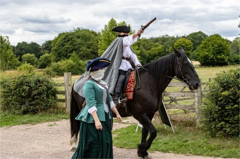 A man dressed as a highwayman rides a dark horse and raises a vintage gun