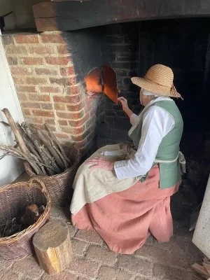 A woman in 18th Century costume sits next to a brick built bread oven in an 18th century cottage