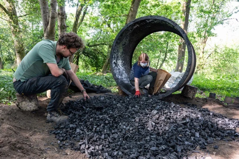 A man and a woman in a woodland are gathering charcoal from a big pile and putting it into bags