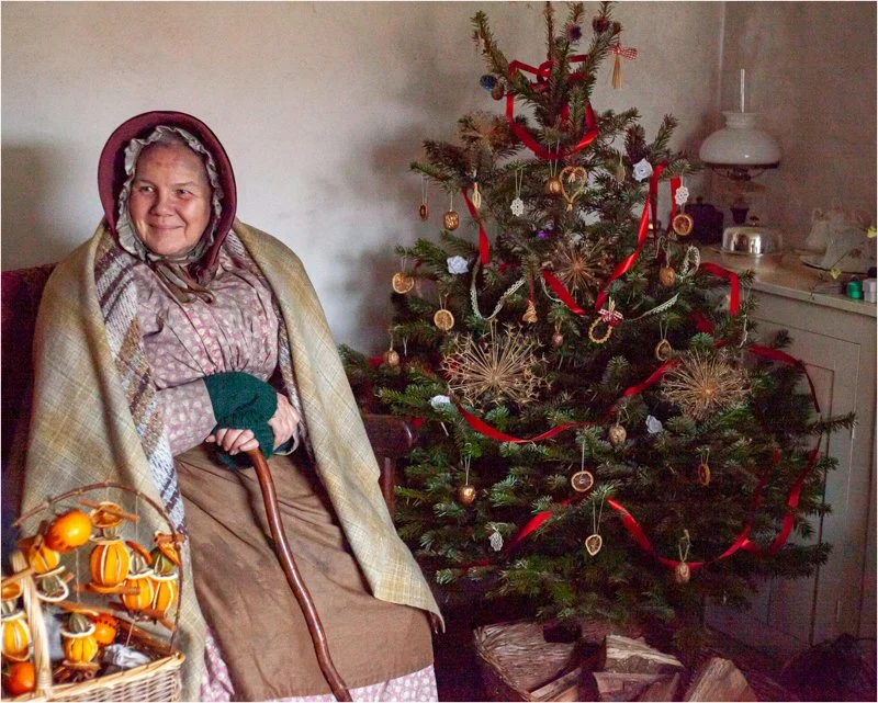 Victorian woman seated beside a traditionally decorated Christmas tree