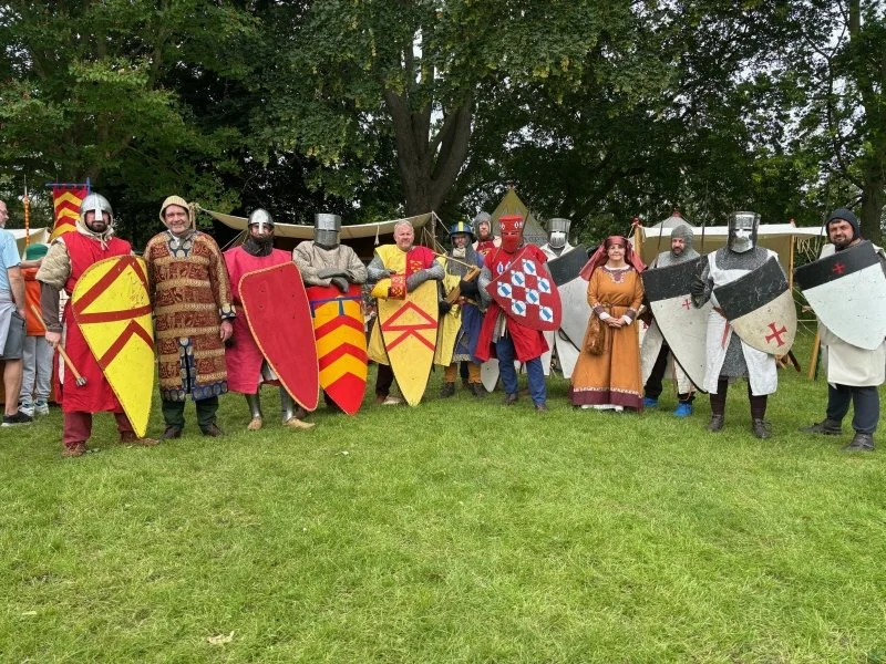 A group of re-enactors dressed as knights stand on grass with trees behind them