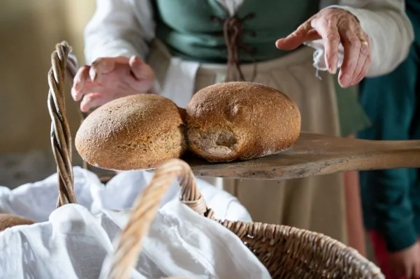 Two cooked round loaves on a wooden board being moved to a basket by a lady in 18th century costume