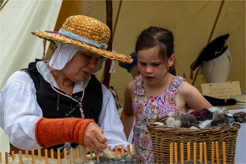 A lady in a traditional outfit and straw hat is weaving on a page loom while a young girl watches