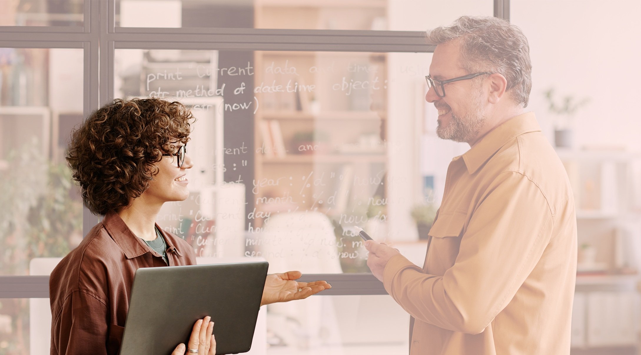 Homem e mulher sorrindo trocando informações dentro de um escritório, com um vidro ao fundo com escrita de código de programação.