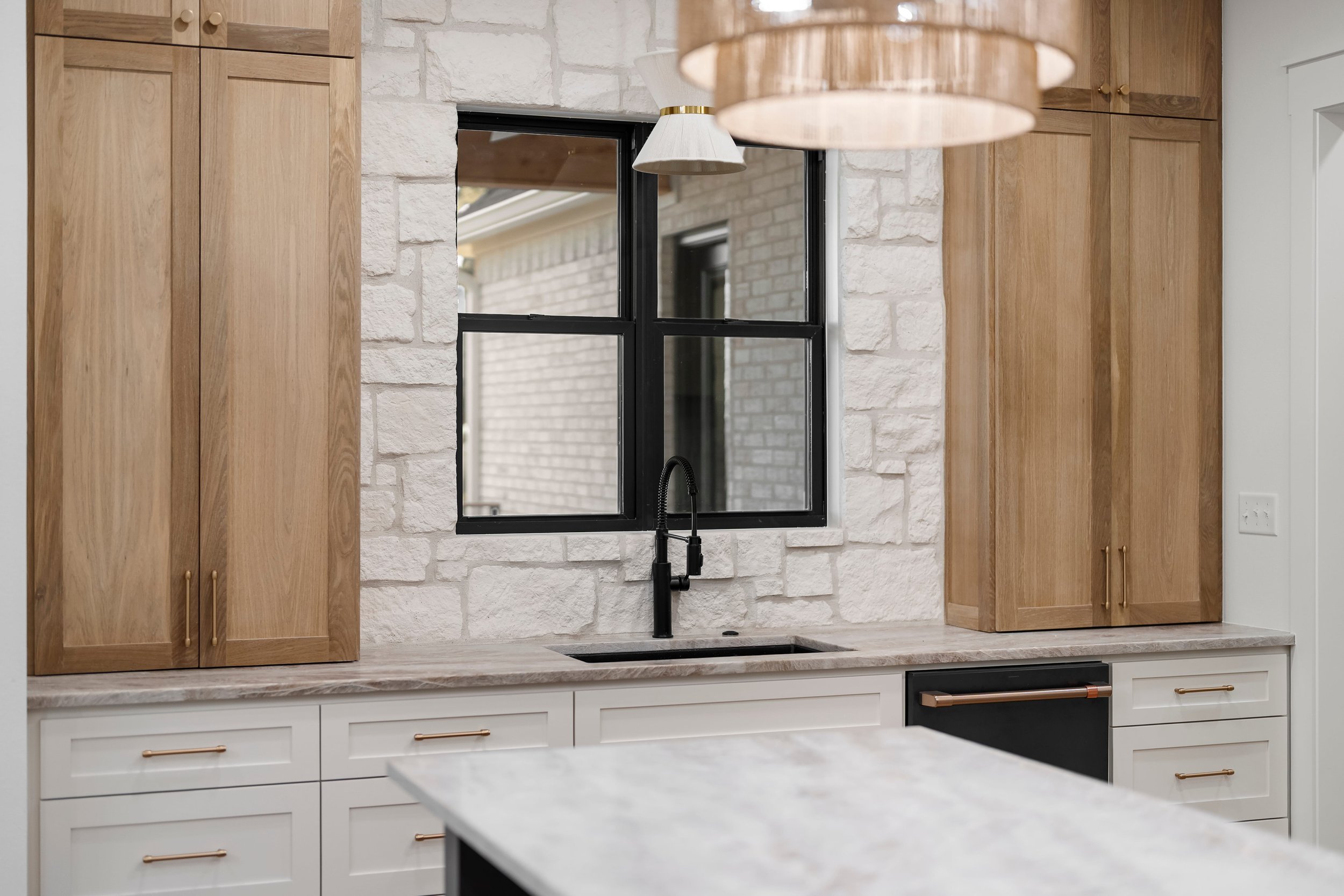 Modern kitchen with white cabinets, gold handles, black faucet, and black-framed window set in light-colored brick wall.