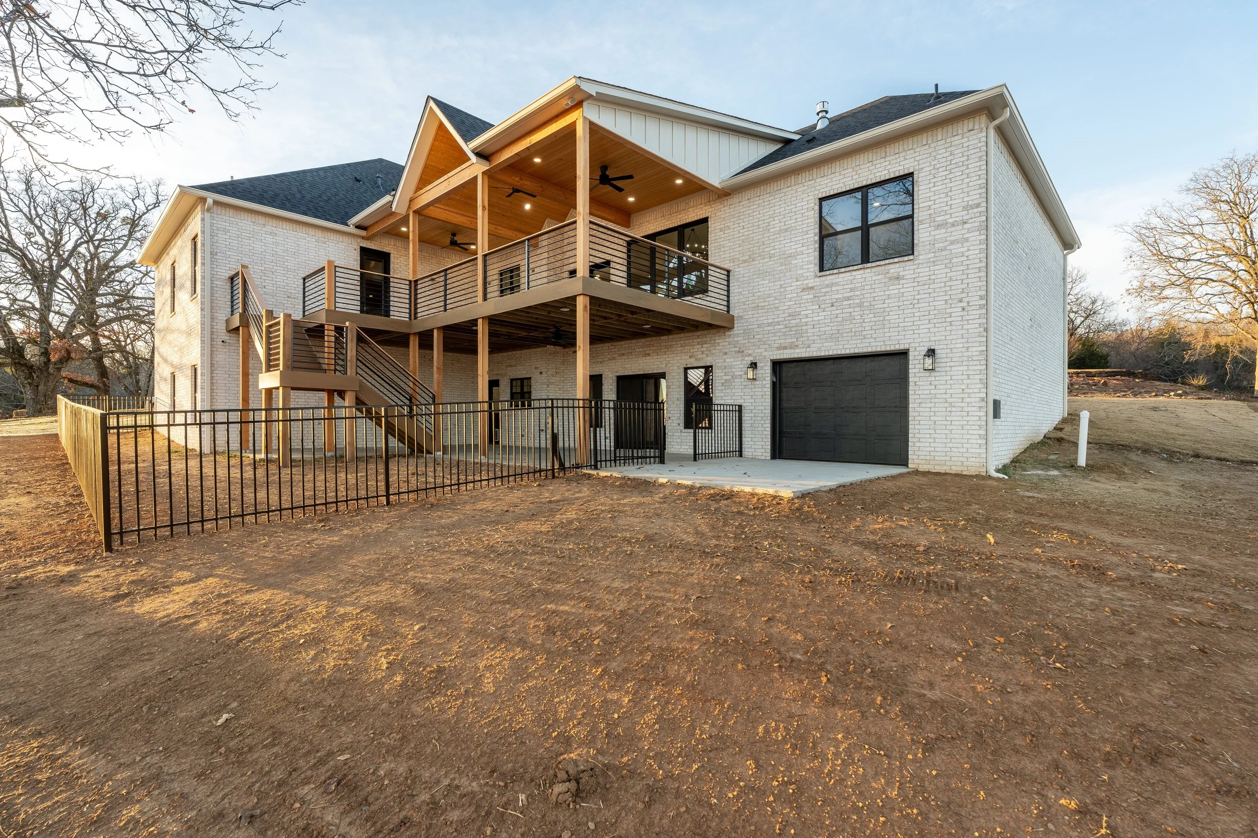 Rear view of a modern two-story house with a large deck, black railing, staircase, black garage door, and white brick exterior, set in a partly wooded area.