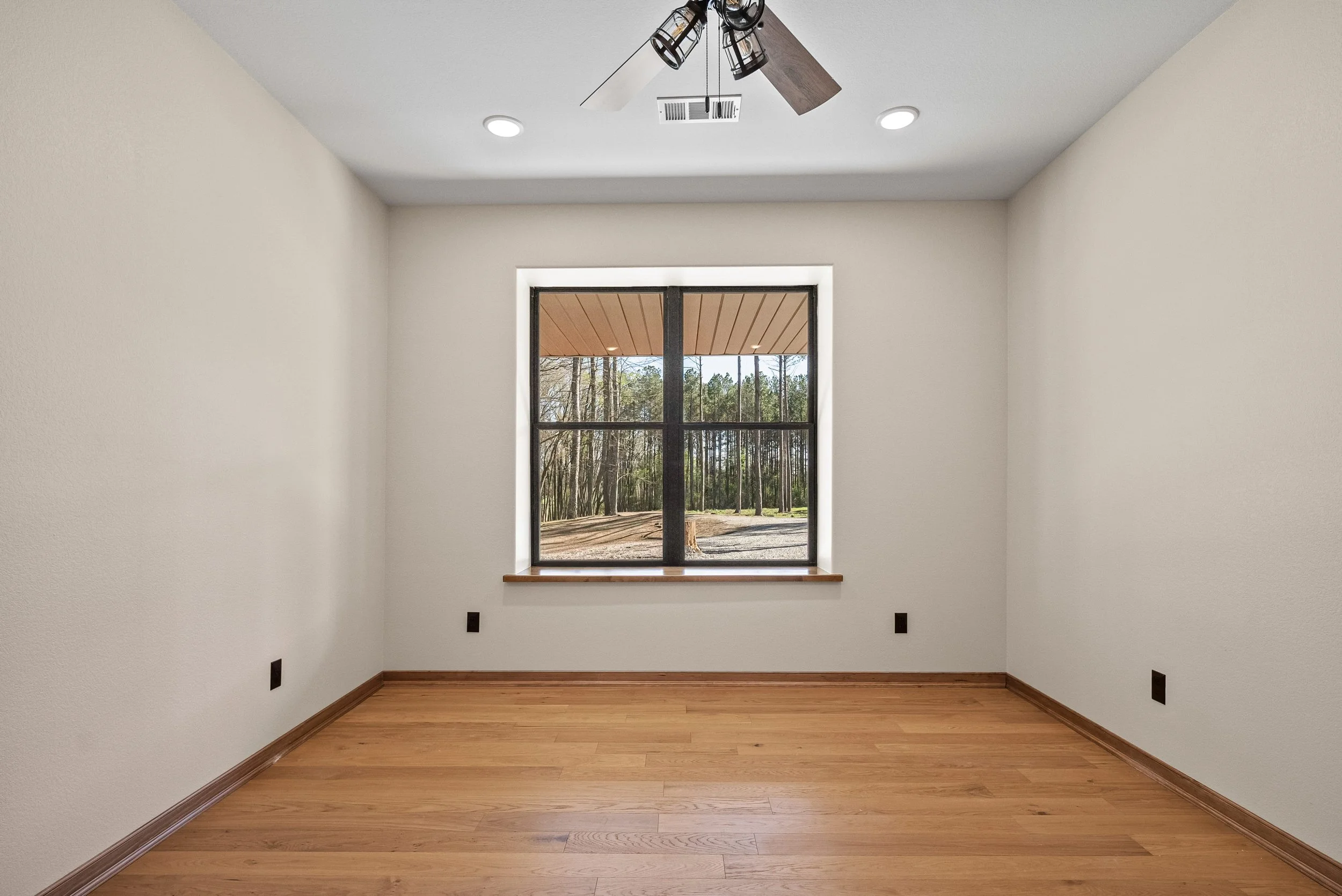 Empty room with white walls, wooden floor, a window looking out to a wooded backyard, and ceiling lights.