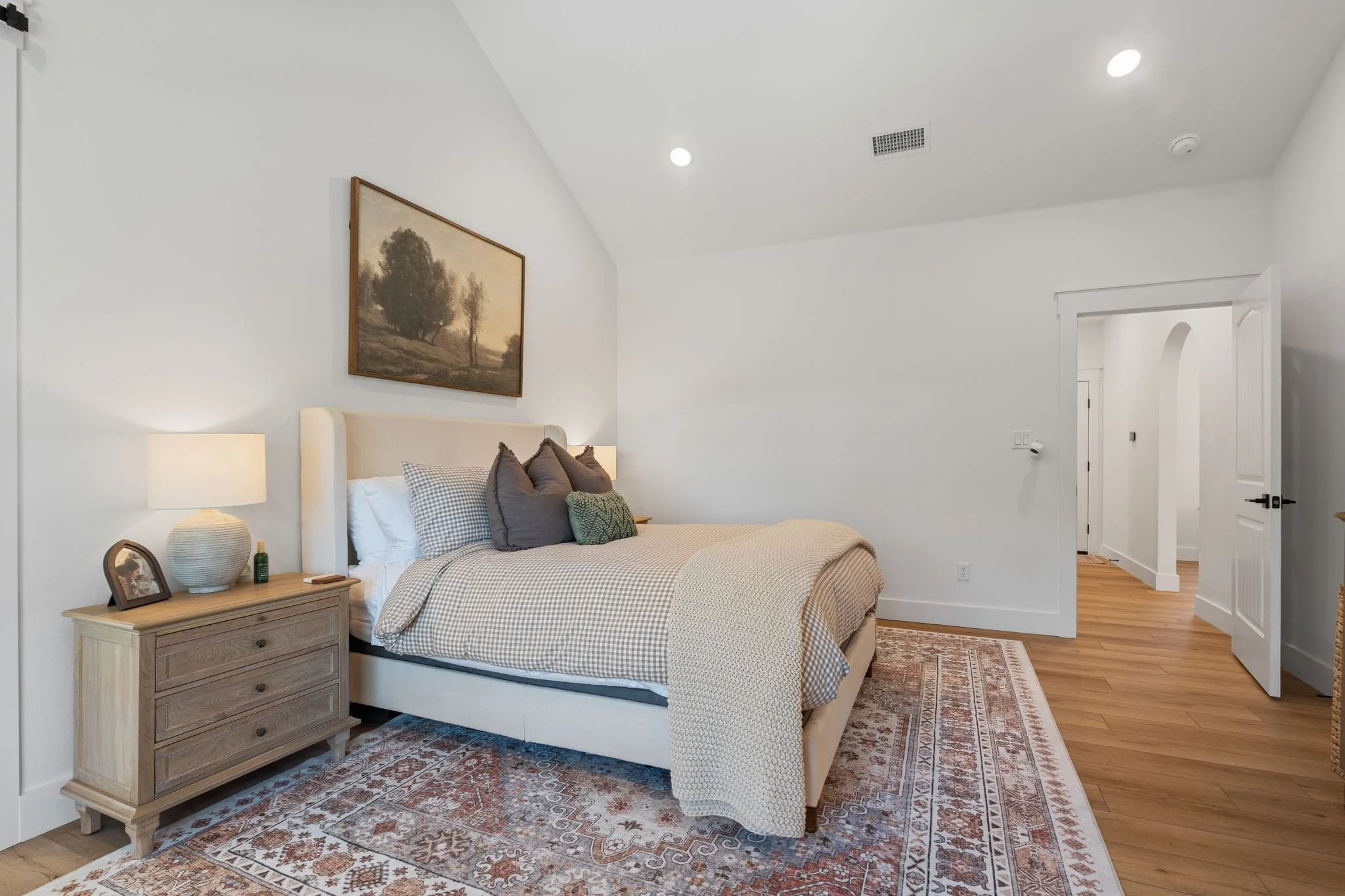 Bright primary bedroom with neutral tones, bedside lamps, textured bedding, and open hallway access.