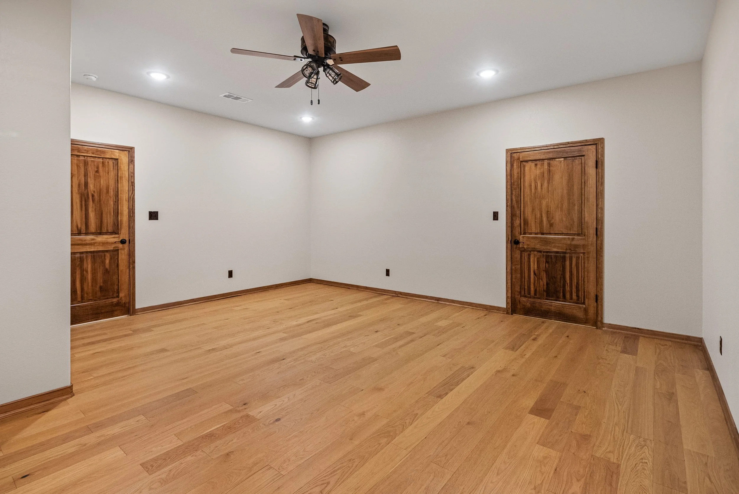 Empty room with hardwood flooring, two wooden doors, white walls, ceiling fans, and recessed lighting.