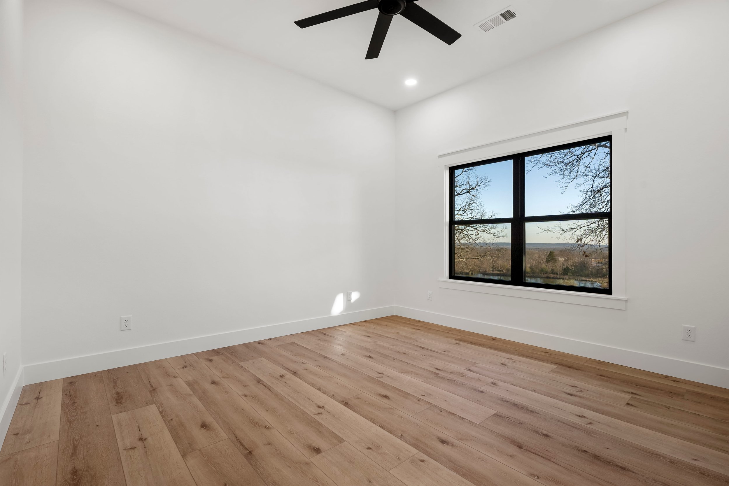 Empty room with white walls, hardwood floors, a window showing an outdoor view, a ceiling fan, and a ceiling light.