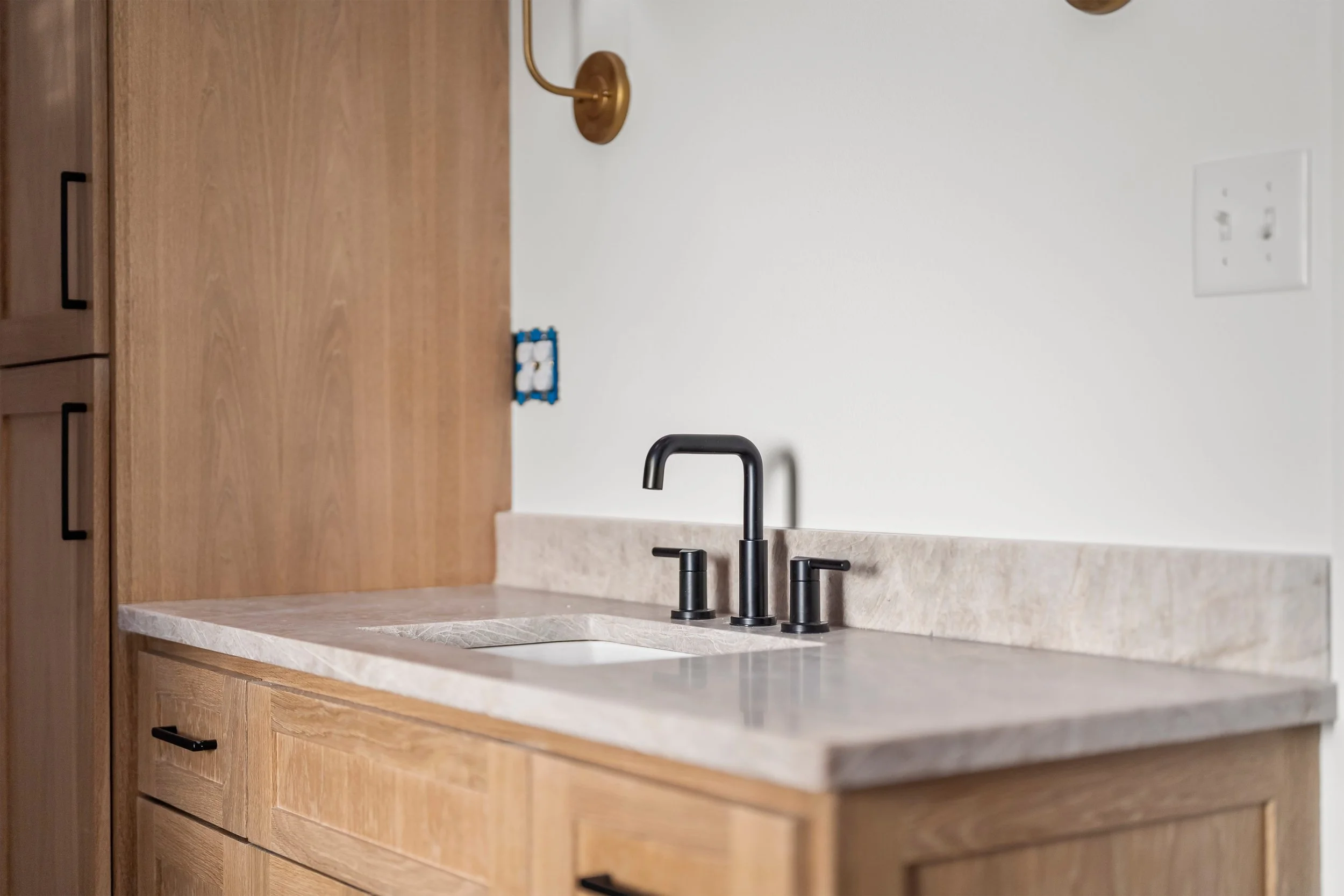A kitchen countertop with a black faucet and sink, wooden cabinets, a wooden wall, a gold wall sconce, an electrical outlet, and a blue and white object on the counter.