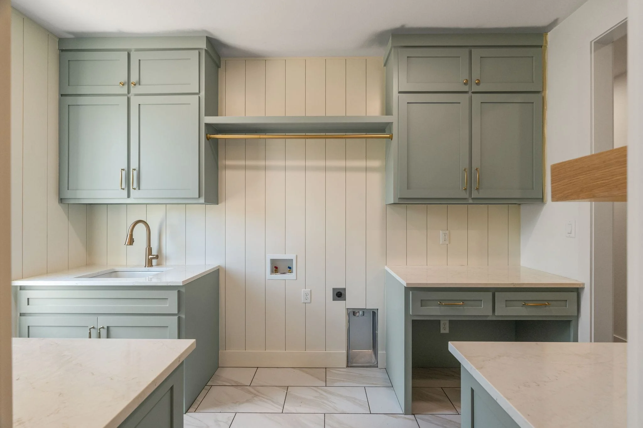 Kitchen with light green cabinets, white countertops, and white paneled walls. Gold handles on the cabinets and a gold rod under the upper cabinets. A sink with a gold faucet is visible below. Electrical outlets and a small compartment on the wall.