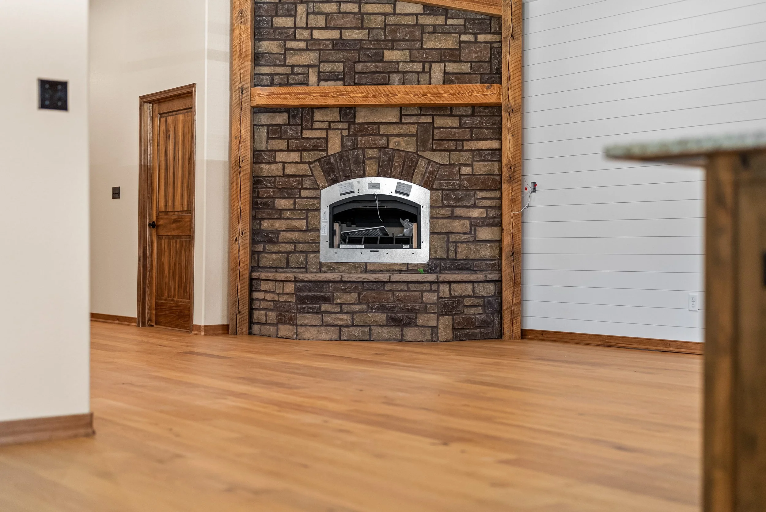 Interior of a room with a brick fireplace, wooden trim, and light-colored hardwood flooring. The fireplace is centered on the wall with a partially installed metal frame. There is a wooden door to the left and a white wall with horizontal paneling to