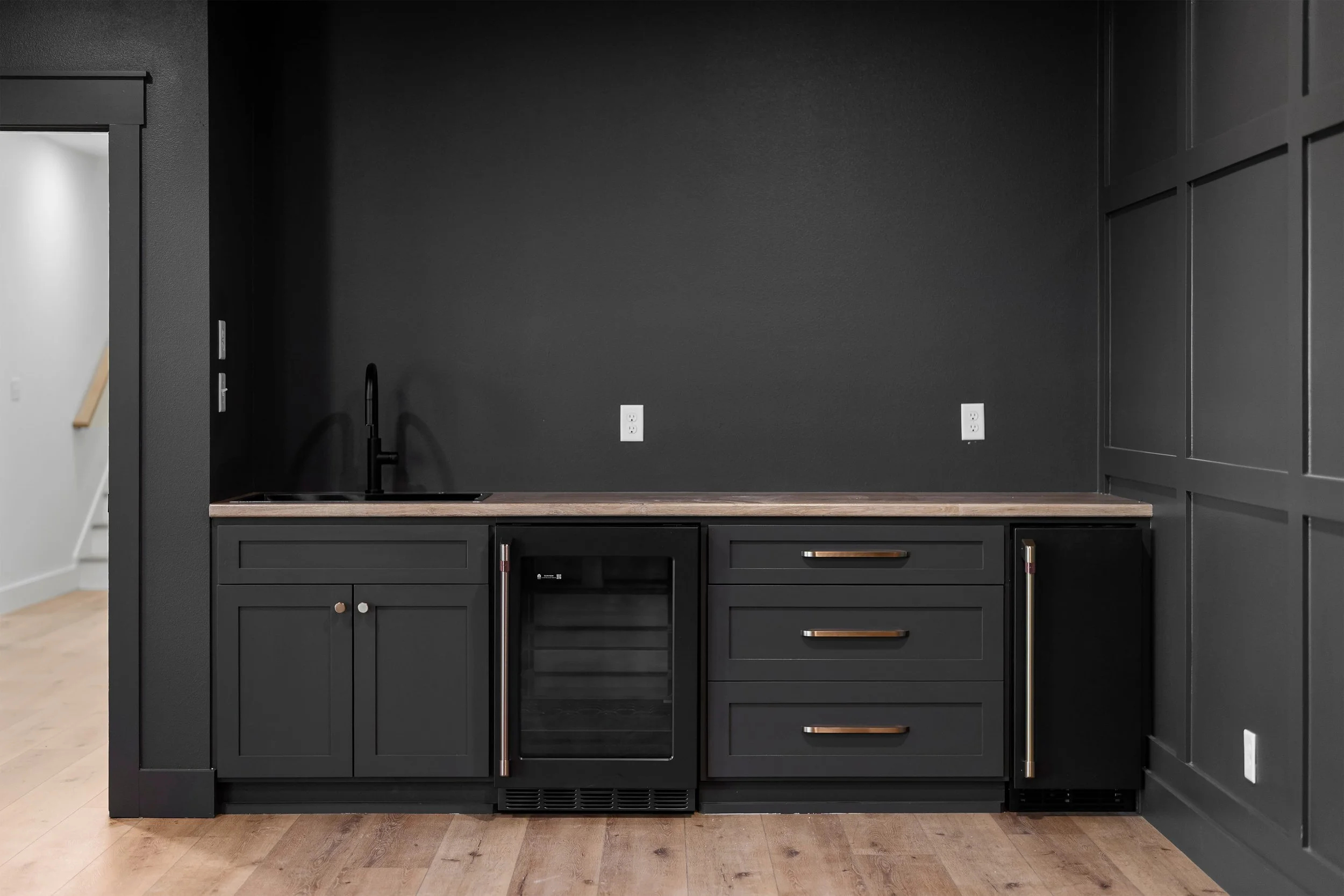 Modern black kitchen with wooden countertop, black cabinet doors and drawers with metallic handles, a small wine fridge, black sink with black faucet, electrical outlets, and hardwood flooring.