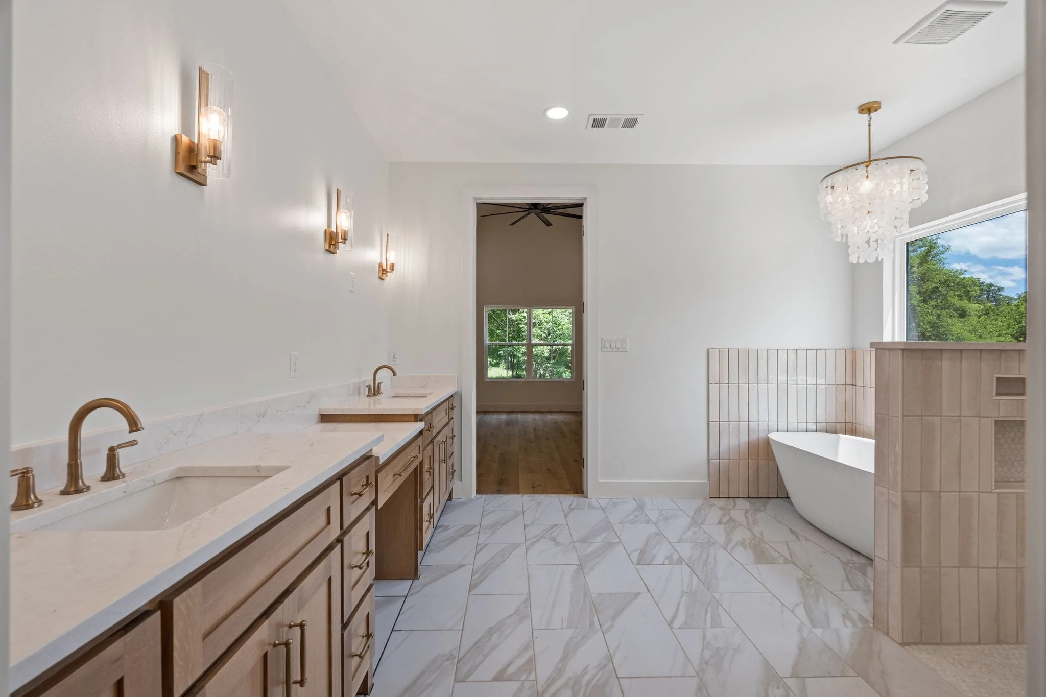 Bathroom with dual sinks, marble countertops, wooden cabinetry, wall-mounted lights, a freestanding bathtub near large window, and wood flooring.