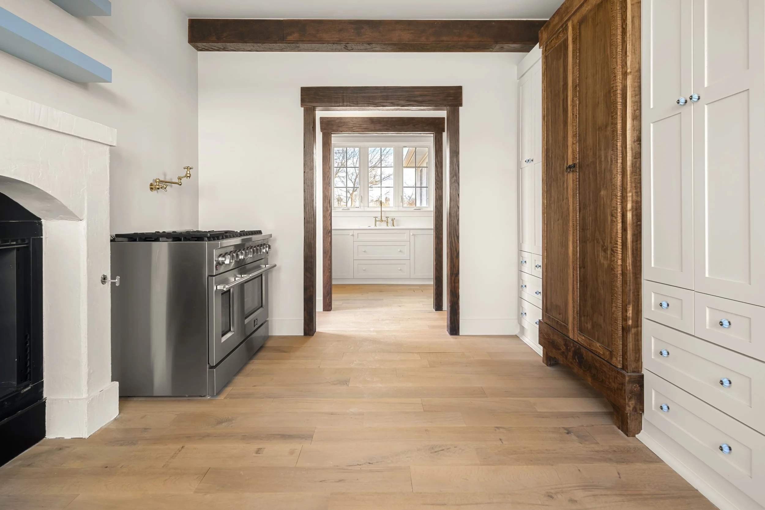 View of a kitchen with wooden floors, white and dark wood cabinetry, a large stainless steel stove, and a doorway leading to a sunny room with a window and sink.