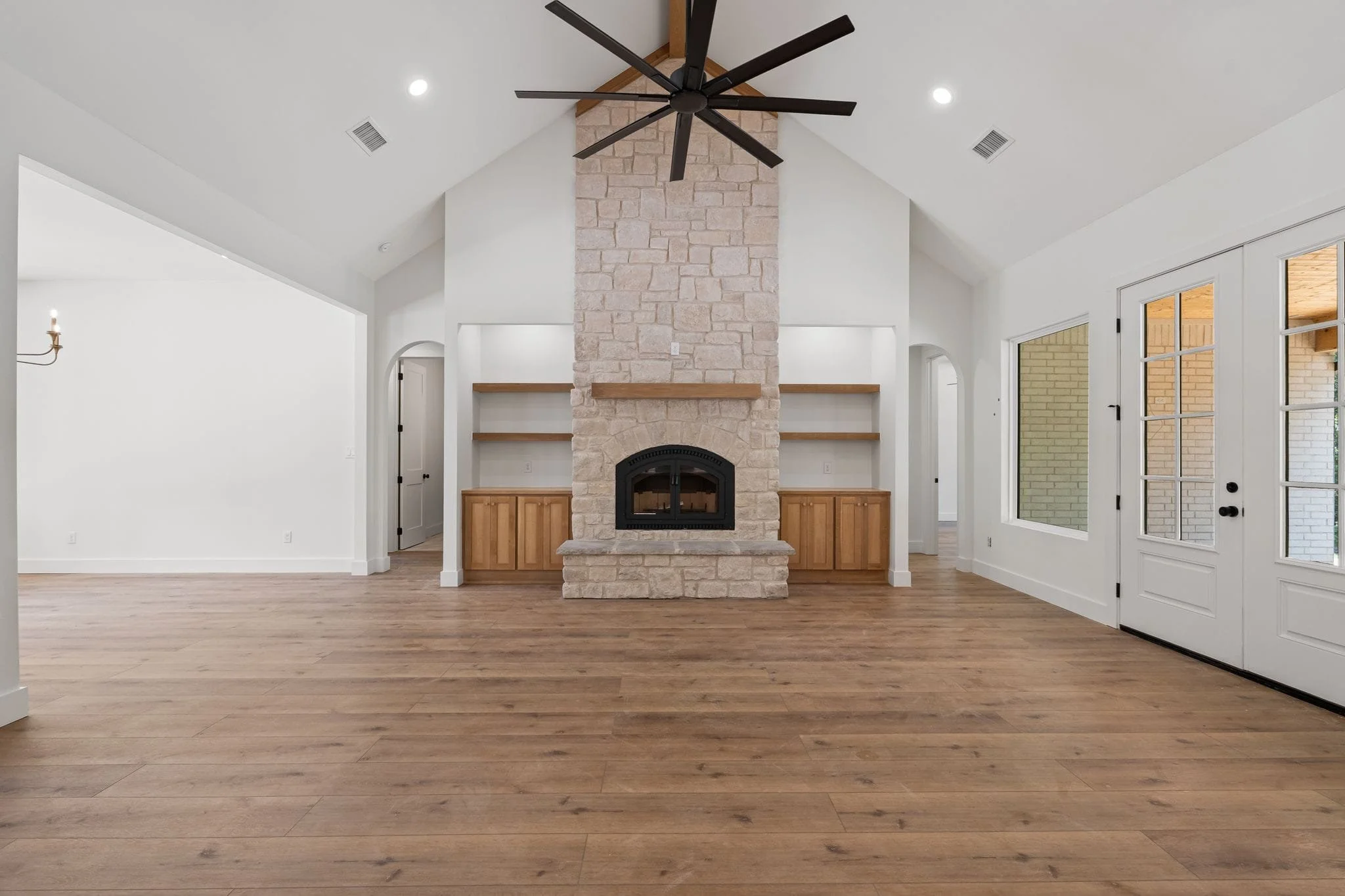 Living room with a large stone fireplace, wooden built-in shelves, and a ceiling fan with black blades. Wooden floors, white walls, and a French door leading outside.