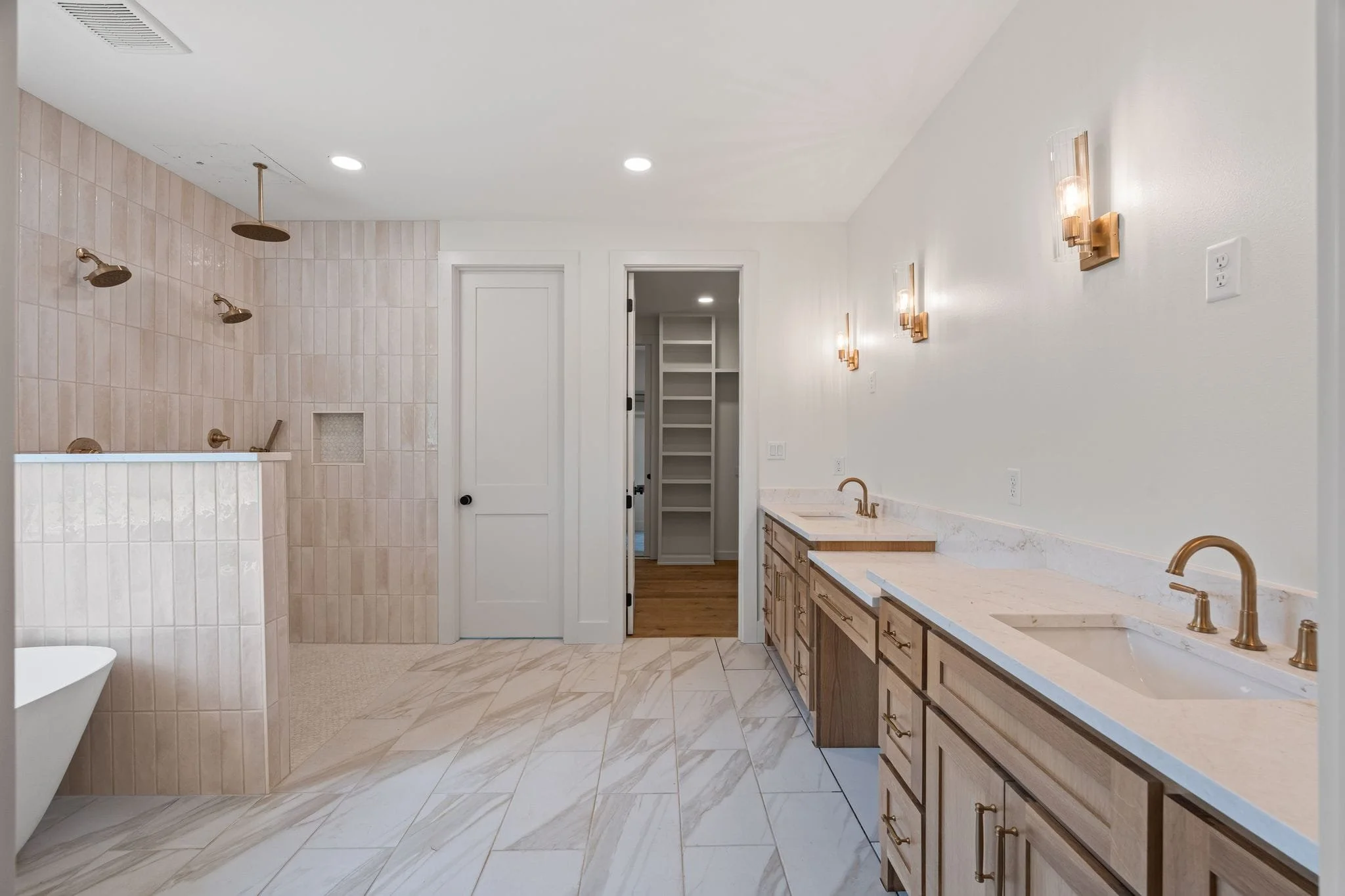 A modern bathroom with a large vanity, wooden cabinets, marble countertops, and brass fixtures. The shower area has beige tiles and multiple rainfall showerheads. The floor is tiled with marble design, and the room is illuminated by recessed ceiling 