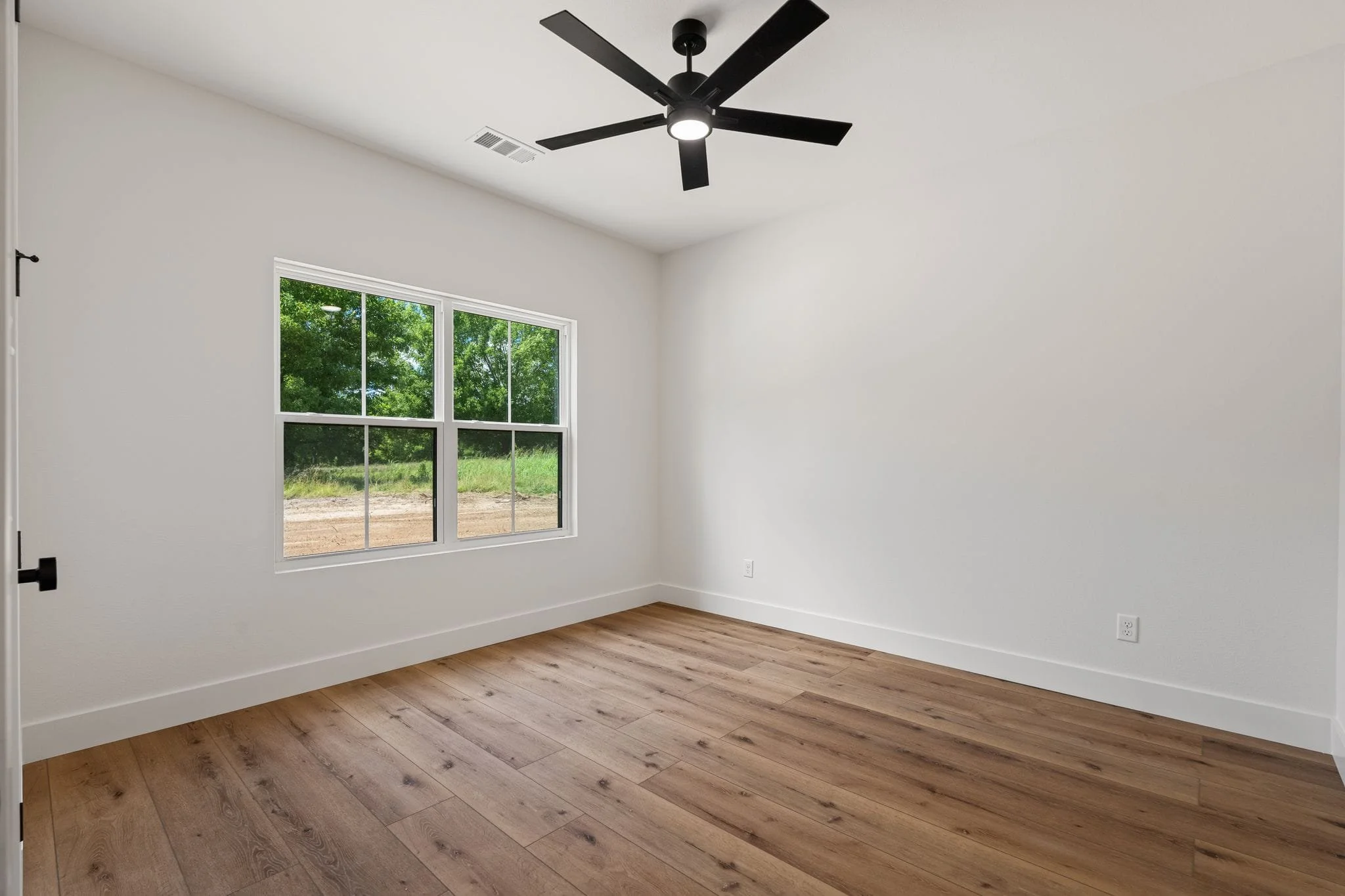 Empty room with white walls, wooden floors, a large window showing greenery outside, and a black ceiling fan.