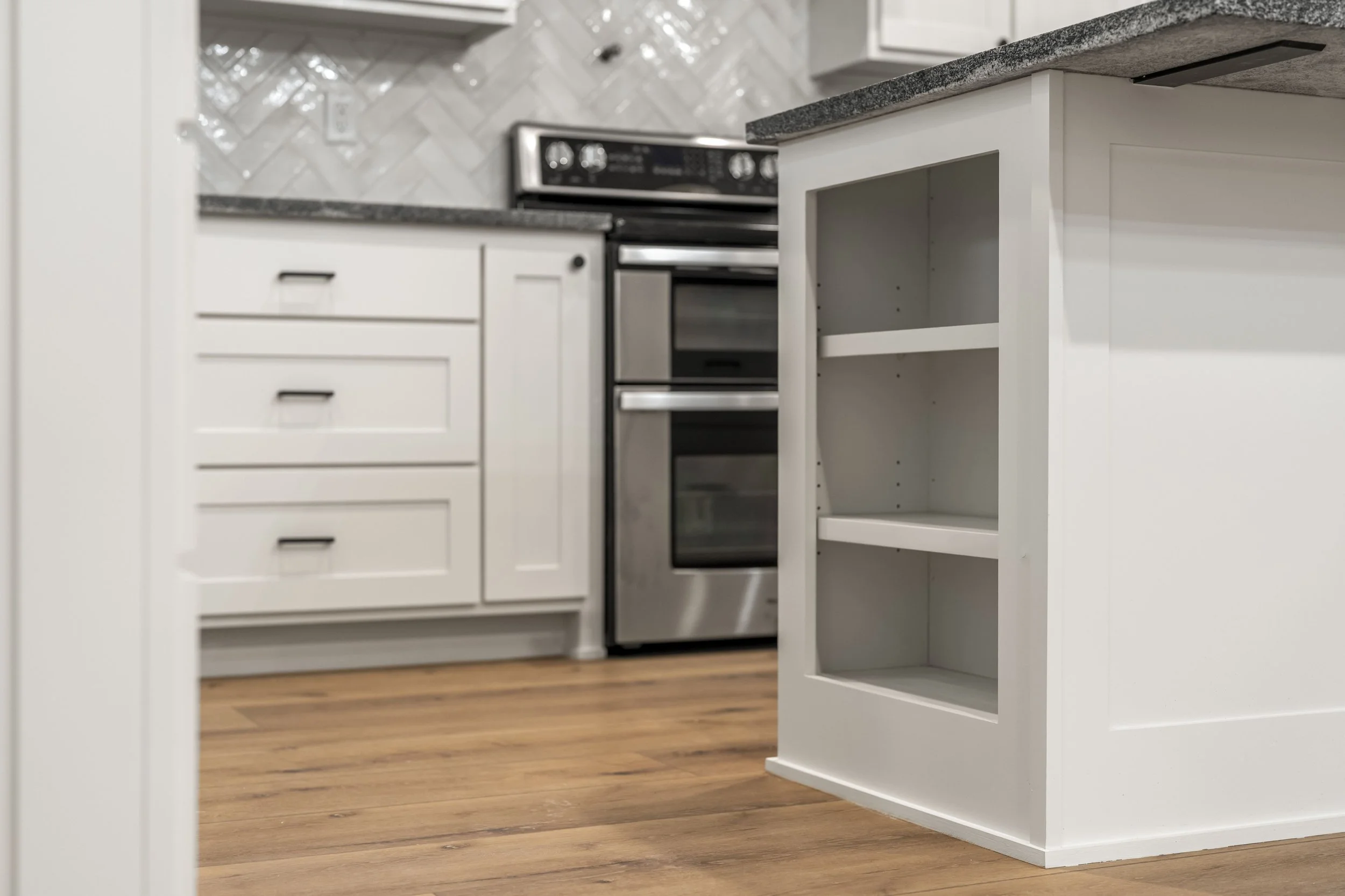 A kitchen with white cabinets, a stainless steel stove, a granite countertop, a wooden floor, and an empty white kitchen island with open shelves.