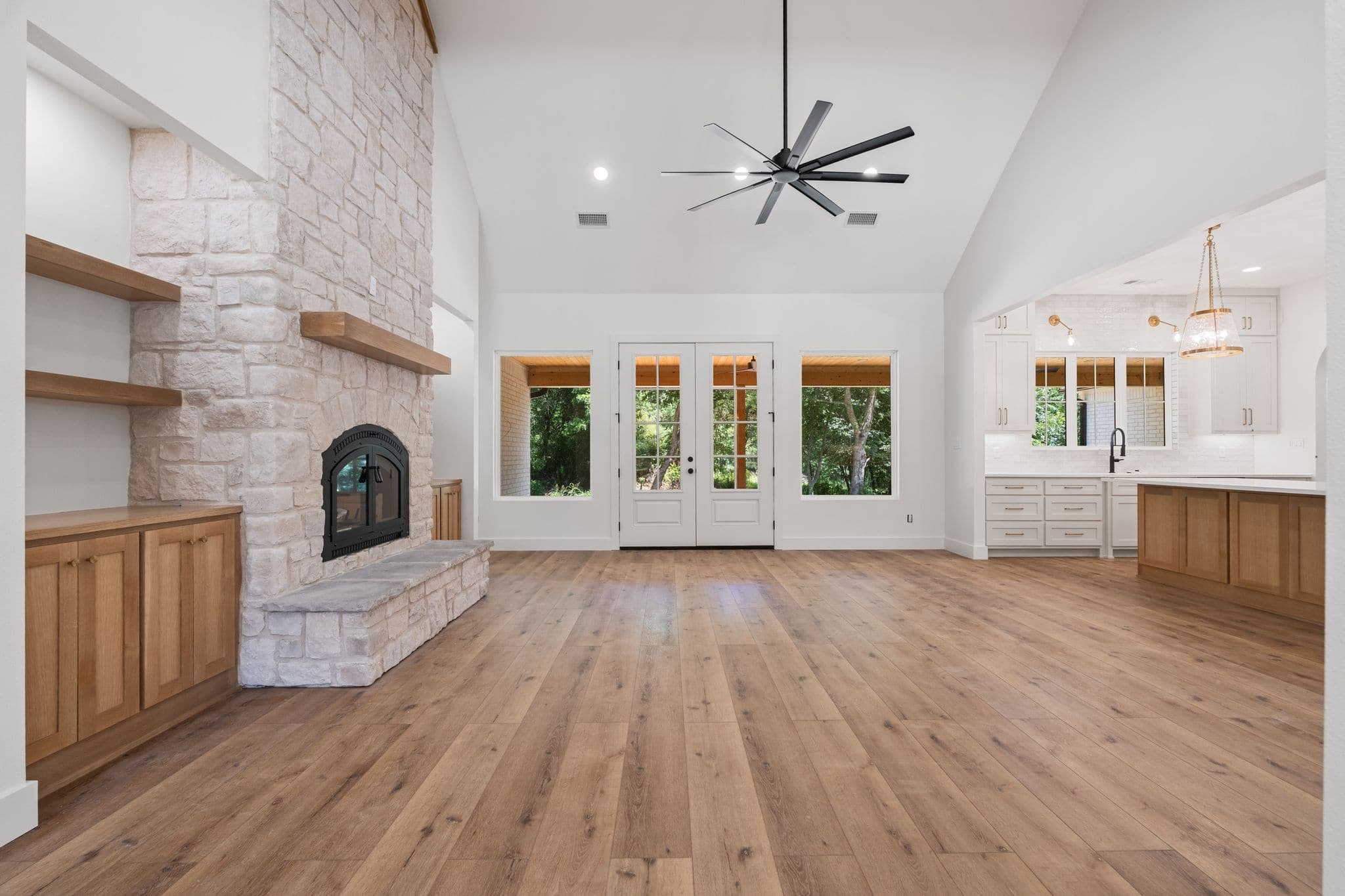 Spacious living area with a stone fireplace, wooden built-in cabinets, hardwood flooring, large windows, and a view of trees outside.