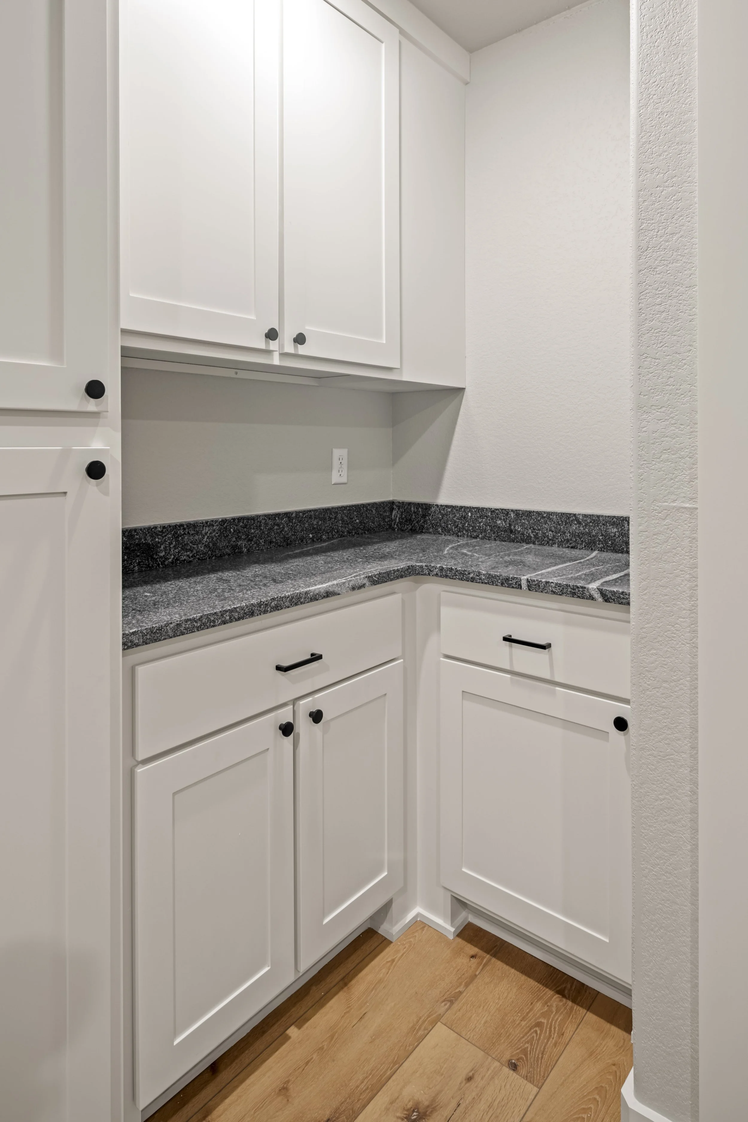 A corner of a kitchen with white cabinets, black knobs, and black granite countertops, with a wooden floor.