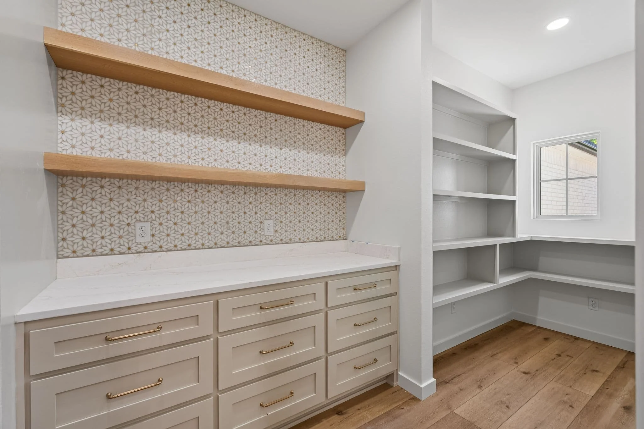 White built-in cabinets with gold handles, open shelving, and a marble countertop. There are three wooden floating shelves on a decorative wallpapered wall with a floral pattern. A window with white trim is visible on the right.