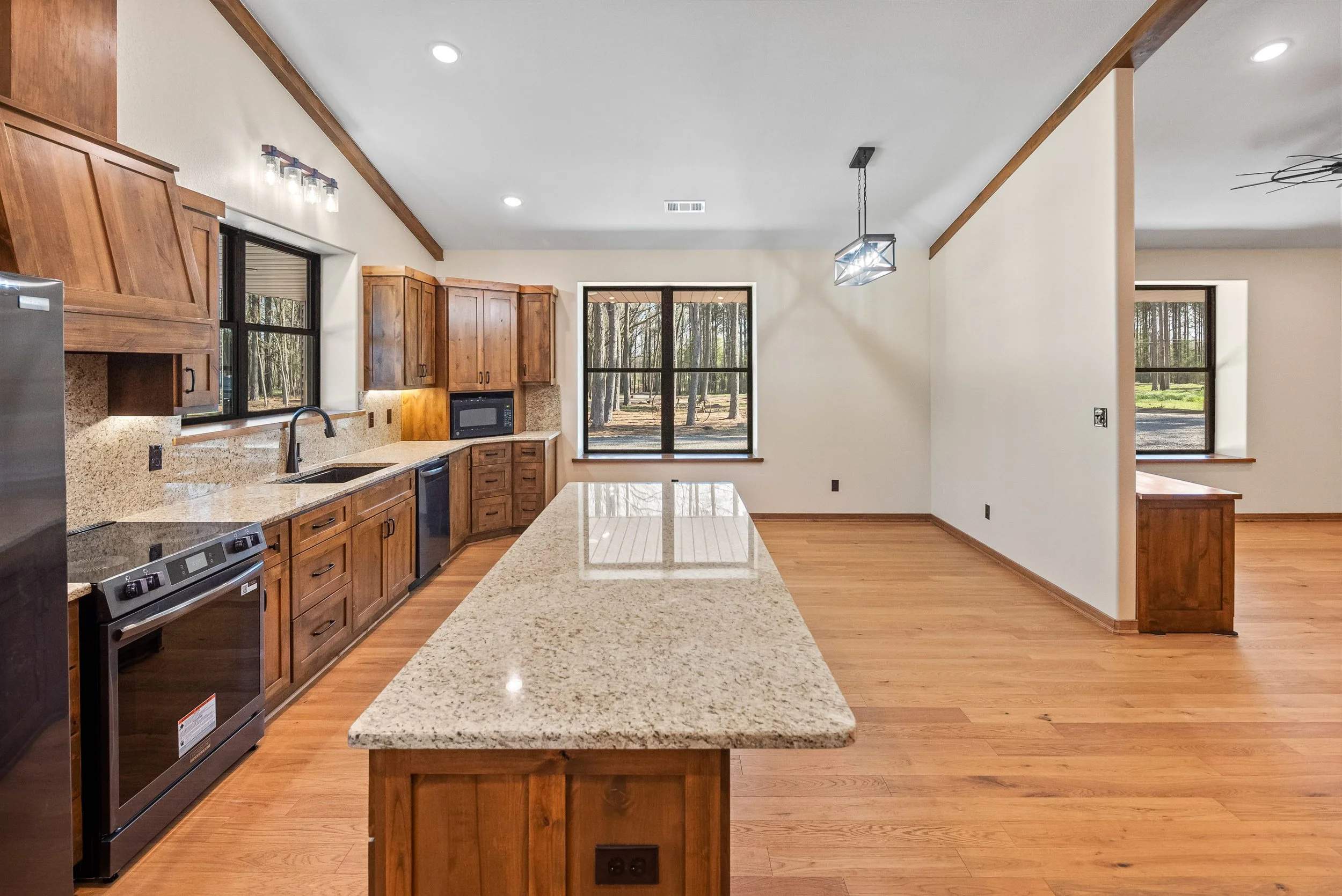 Empty kitchen with wooden cabinets, granite countertops, black appliances, and large windows showing trees outside.