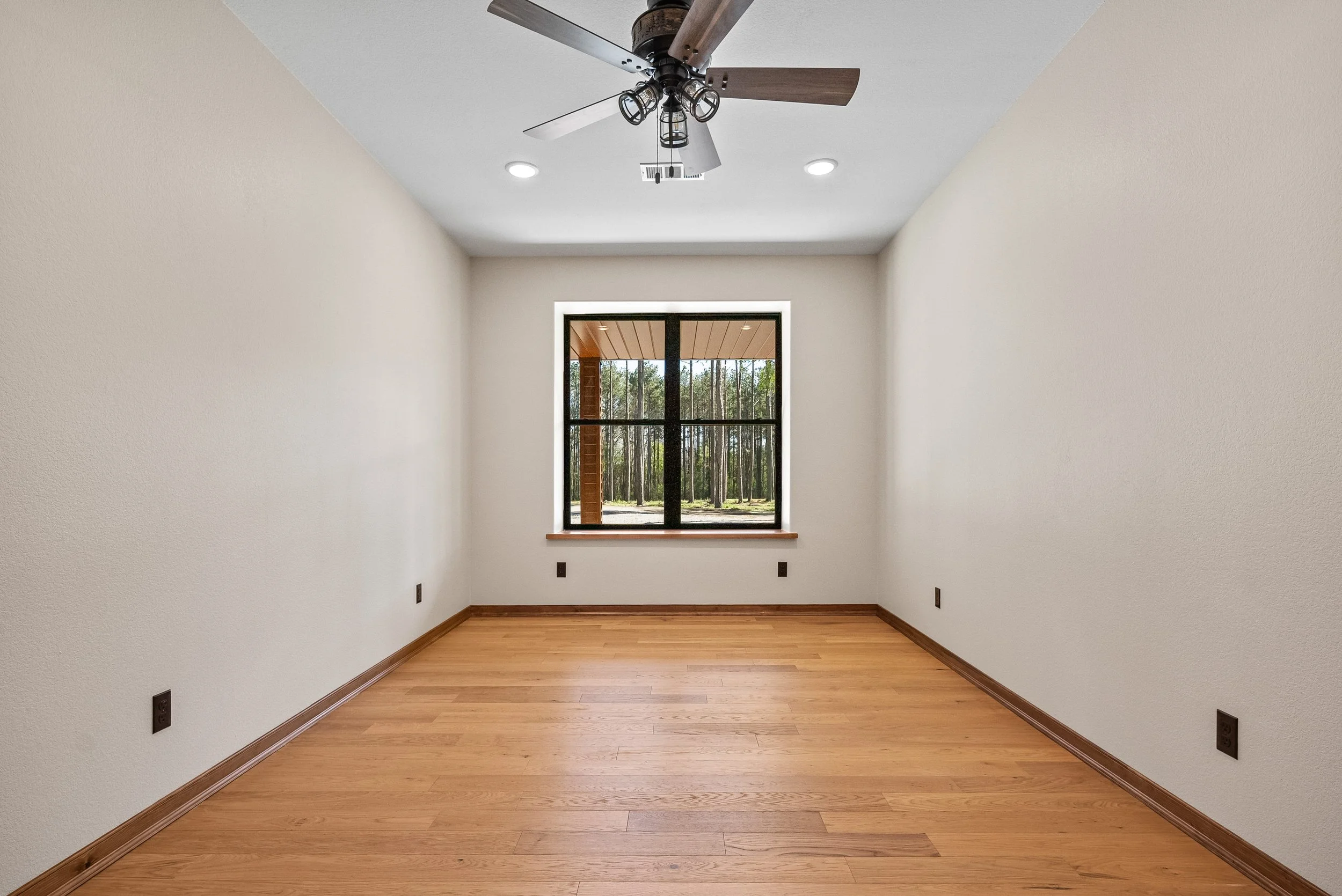 Empty room with wood flooring, white walls, a window showing a view of trees, a ceiling fan, and recessed lighting.