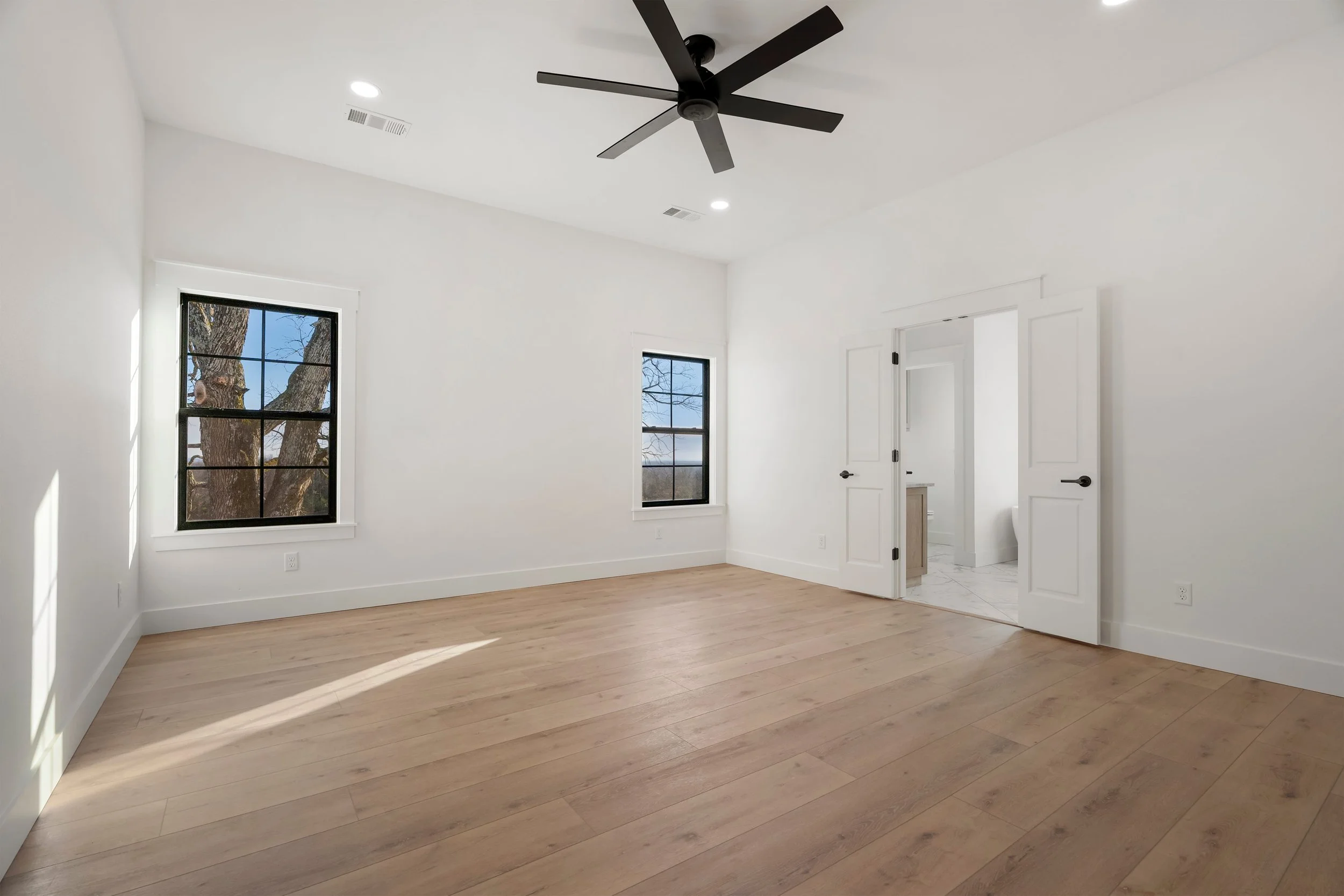 Empty white bedroom with two black-framed windows, light wood flooring, a black ceiling fan, and an open door leading to a bathroom with tile flooring.