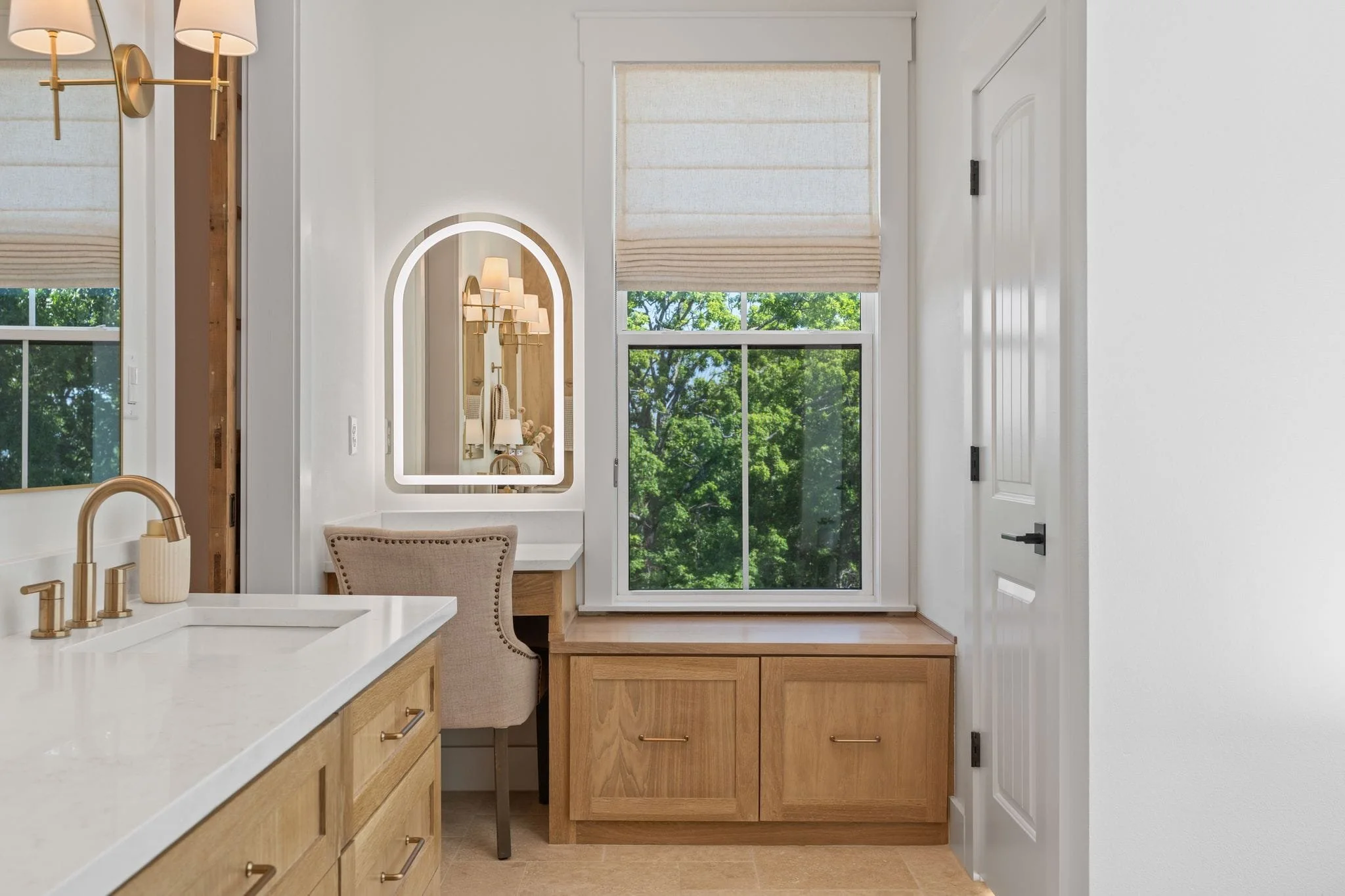 Bathroom vanity area with built-in makeup station, illuminated mirror, window bench seating, and natural wood cabinetry.