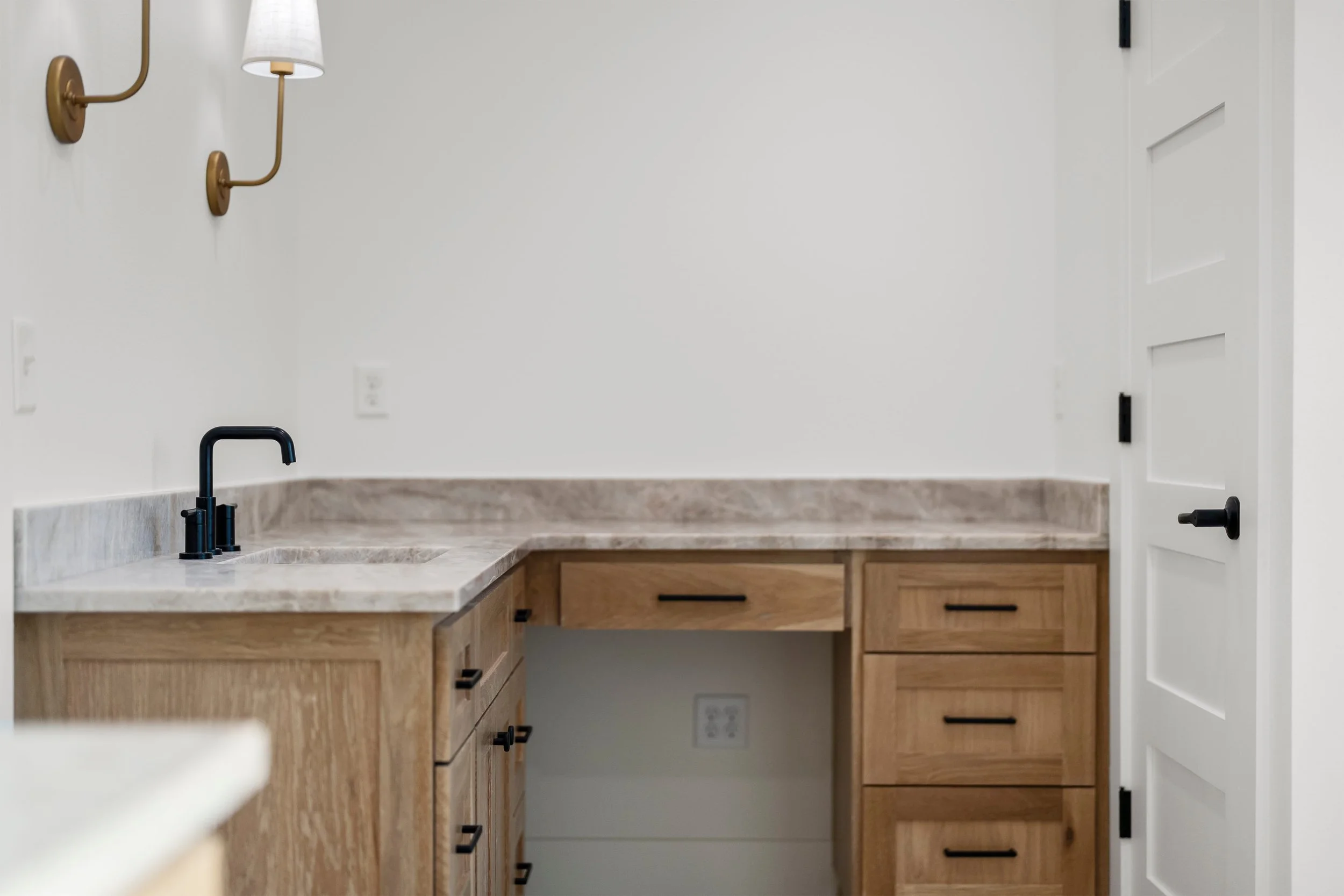 Empty kitchen counter with wooden cabinets, black faucet, and white walls with wall sconces.