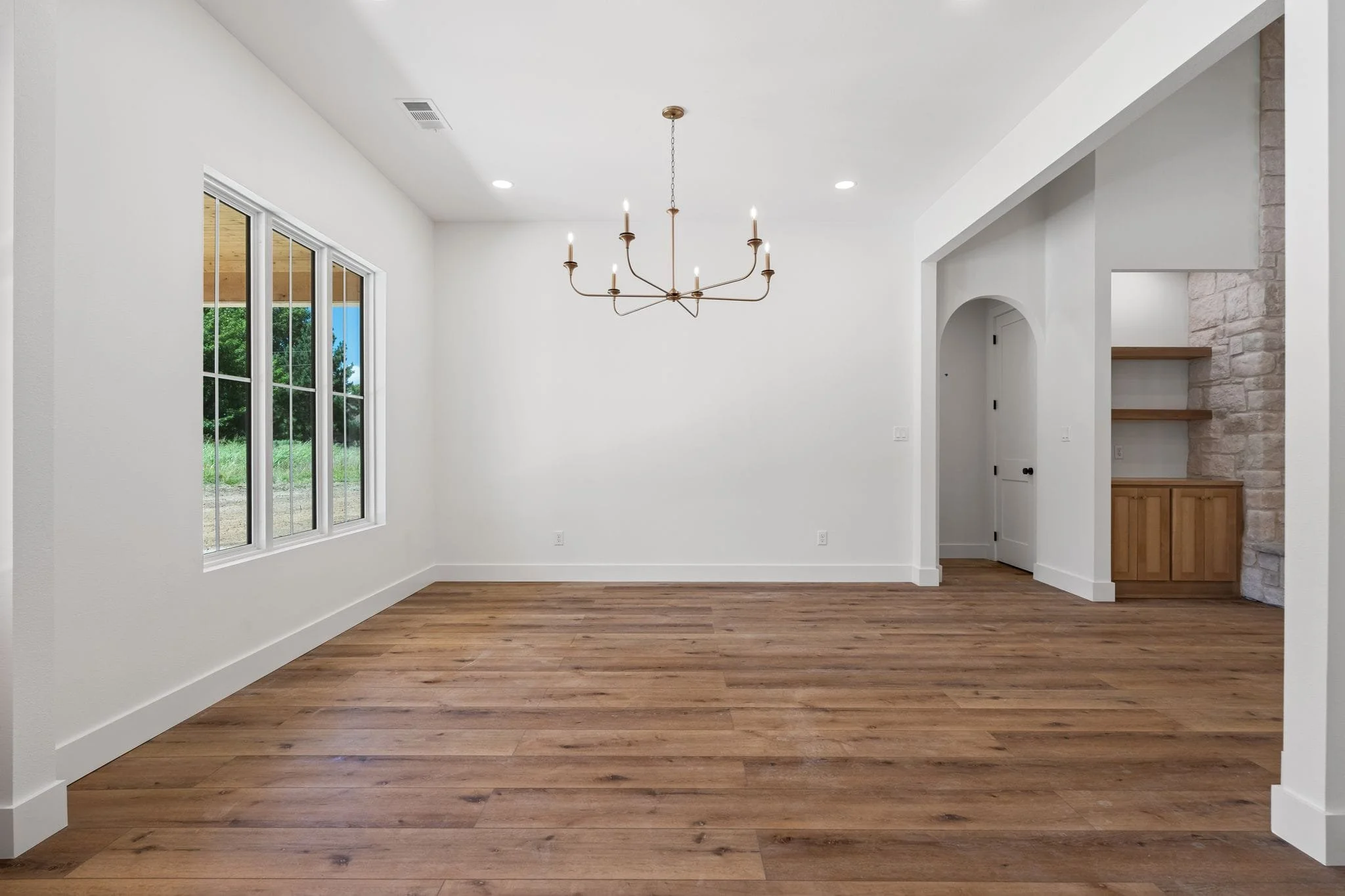 Empty room with white walls, large window, wooden flooring, and a chandelier hanging from the ceiling.