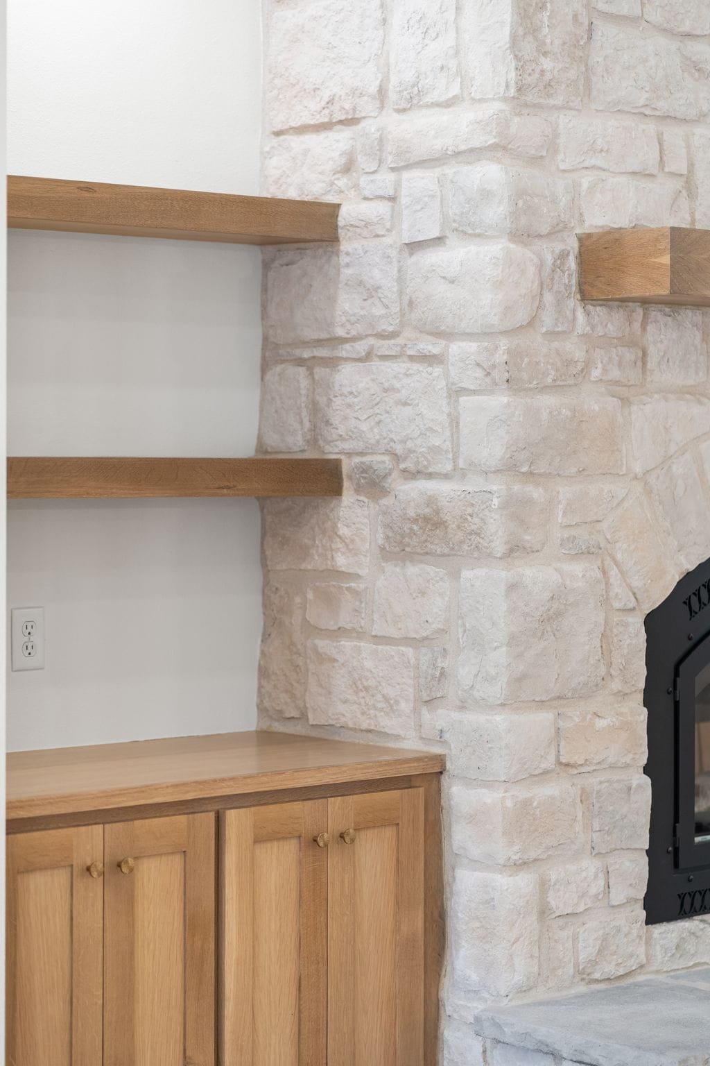 A corner of a room with wooden shelves and a wooden cabinet against a white brick wall, with a black fireplace on the right.
