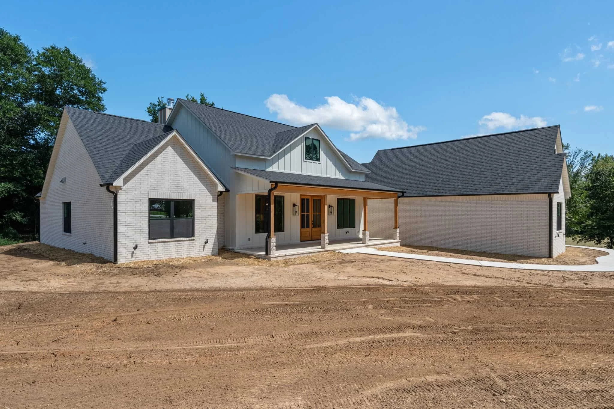 A newly built white brick house with a dark gray roof, front porch with wooden door, surrounded by bare soil, with green trees and a blue sky in the background.