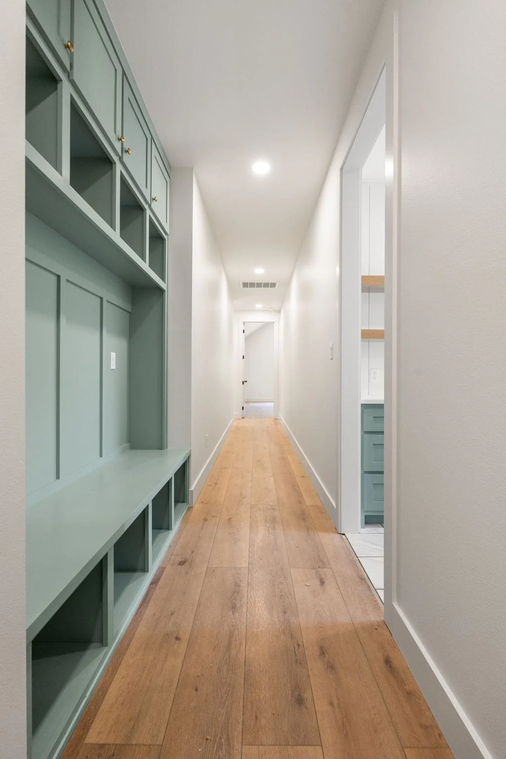 Quiet hallway with white walls, wooden floor, built-in green shelving on the left, and a doorway on the right leading to a space with white cabinetry and wooden shelves.