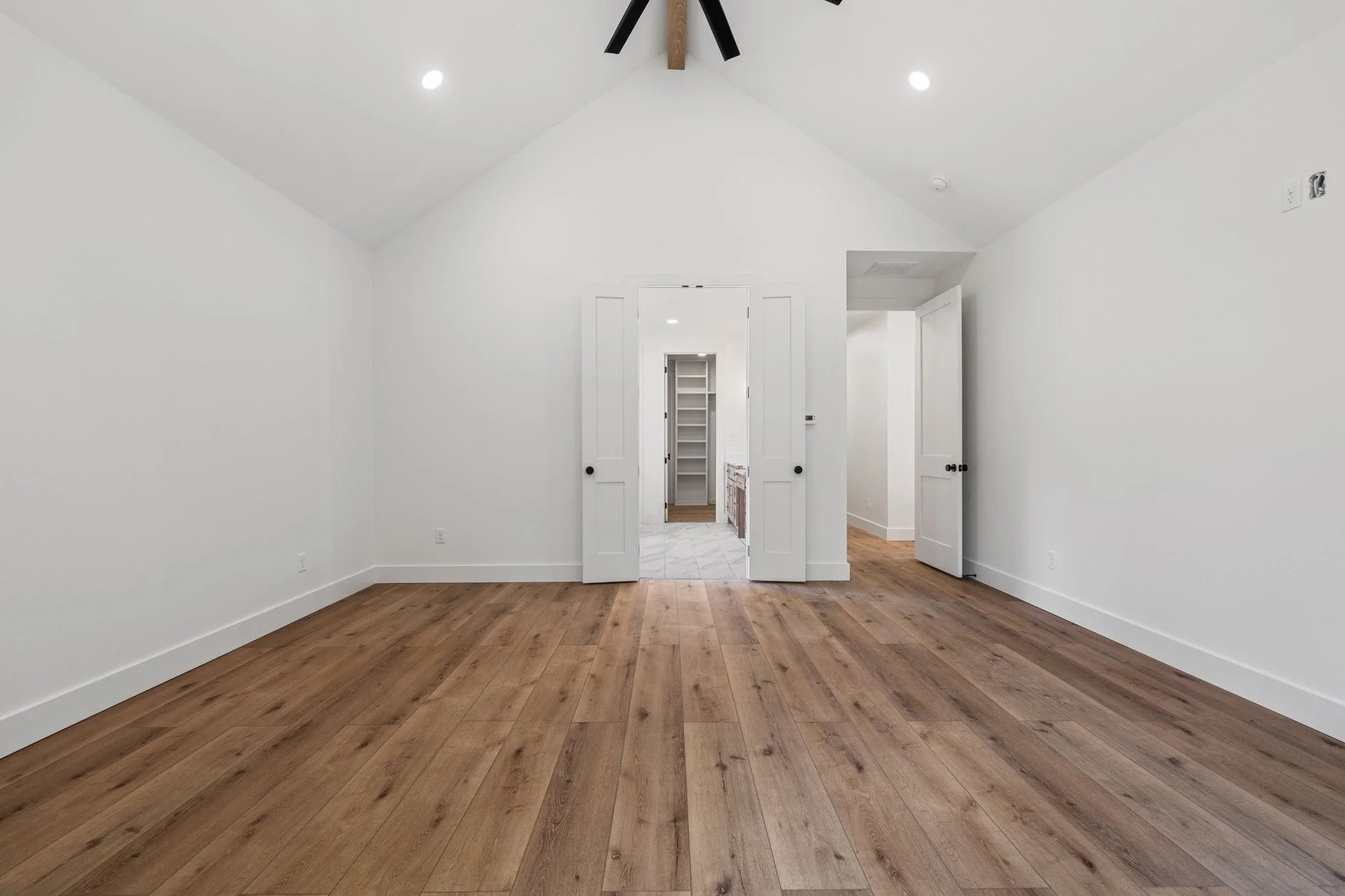 A room with white walls, ceiling, and trim, featuring hardwood flooring and two open white doorways leading to different rooms; ceiling has two recessed lights and a black and brown ceiling fan.