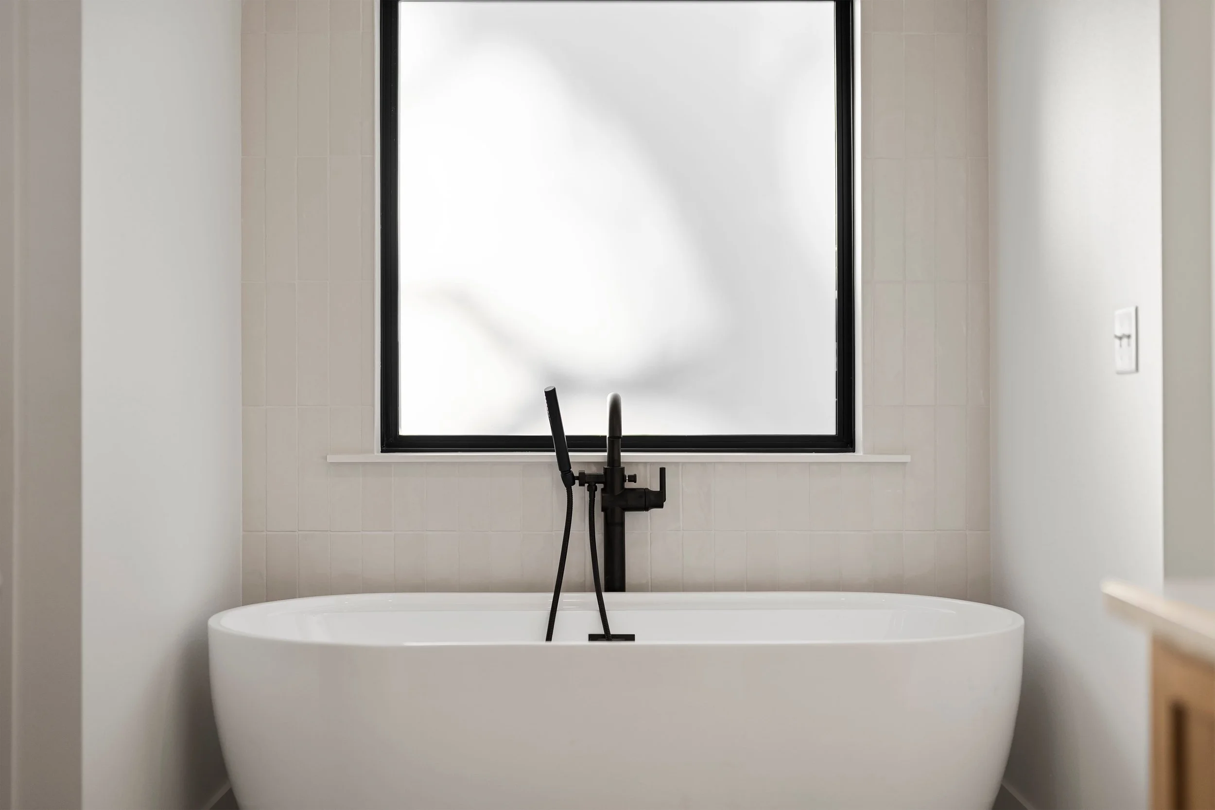 Modern bathroom with a white freestanding bathtub positioned beneath a large frosted window, with a black faucet and handheld showerhead.